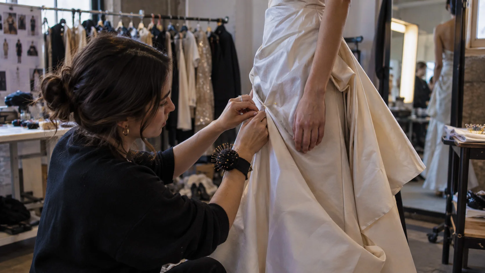A stylist pins the hem of a garment while the model stands still on a fitting block in a studio wardrobe area.